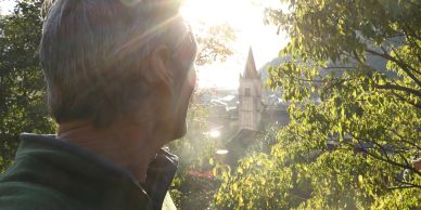 Man looking at a sunlit church tower through trees.
