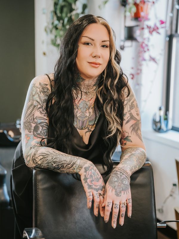 A tattooed woman with long black and blonde hair leans on a chair, smiling confidently.