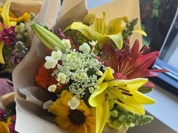 A vibrant bouquet of yellow lilies, sunflowers, and other colorful flowers wrapped in beige paper.