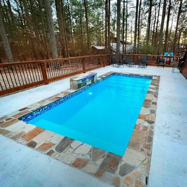 A rectangular swimming pool surrounded by stone tiles and a wooden fence in a forested backyard.