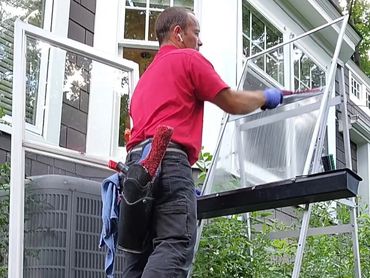 Man cleaning window screens outside a house with tools and gloves.