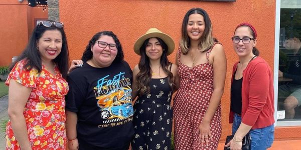 Five smiling women standing together against an orange wall.