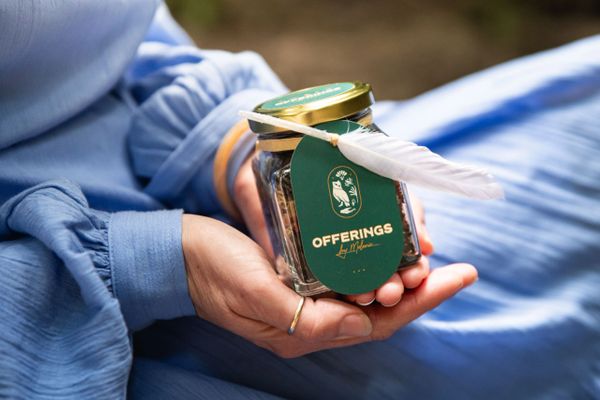 A person in blue holding a jar labeled 'OFFERINGS' with a feather attached.