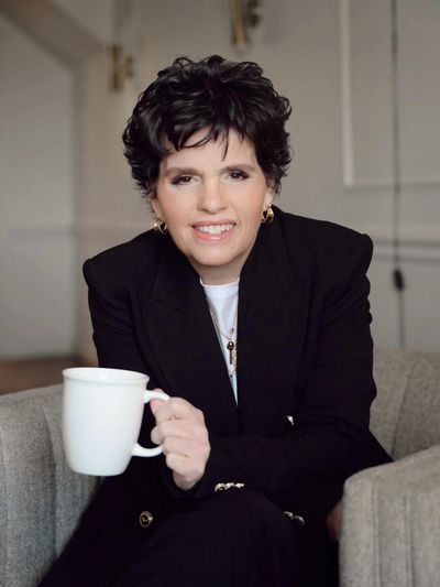 Smiling person with short curly hair holding a white mug, dressed in a black blazer.