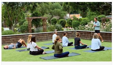 Picture of several people seated on yoga mats in a garden and instructor standing in front.