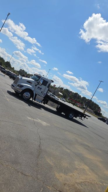 Flatbed tow truck parked in a sunny, spacious parking lot under a blue sky.