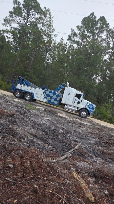 A blue and white tow truck parked on a dirt road beside a forest.