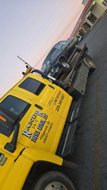 Yellow tow truck from RANDR Diesel Repair carrying a dark classic car at sunset.