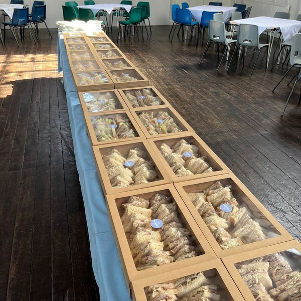 Boxes of sandwiches arranged on a long table in a room with chairs and tables.
