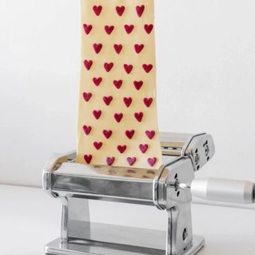 Hand holding pasta dough embedded with small red heart shapes over a pasta machine.