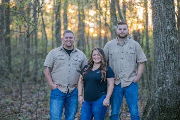 Three people standing in a forest during sunset, smiling at the camera.