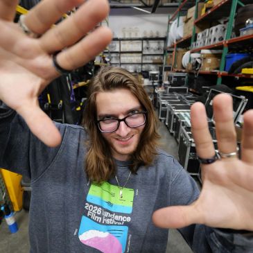 Young man with glasses playfully framing the camera with his hands in a workshop.