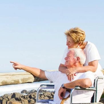 Man in a wheelchair pointing to the ocean as a woman wraps her arms around him.