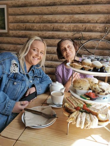 Two women enjoying afternoon tea with sandwiches and desserts in a cozy wooden cabin.