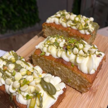 Three slices of moist cake with white frosting, pistachios, and drizzle on a wooden board.