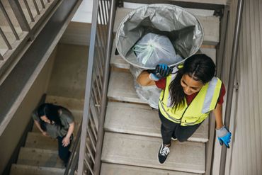 Woman in safety vest carrying trash bag up stairs, another person climbing below.
