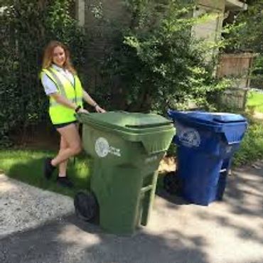 A woman in a safety vest stands next to green and blue recycling bins outdoors.