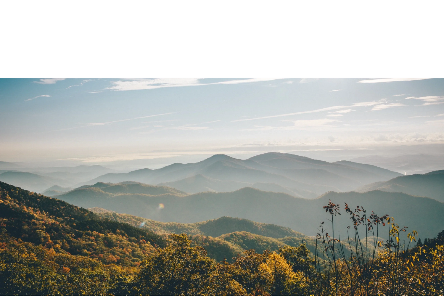 Sunlit mountain ranges with autumn foliage and misty valleys under a clear sky.
