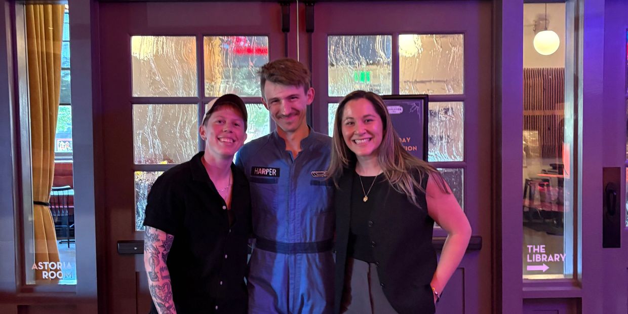 Three friends smiling together in front of glass doors at a cozy venue with signs for Astoria Room and The Library.