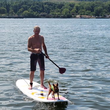 Man paddleboarding with a dog wearing a life jacket on a lake.