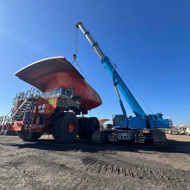 A blue crane lifts the bed of a large red mining truck under clear blue sky.