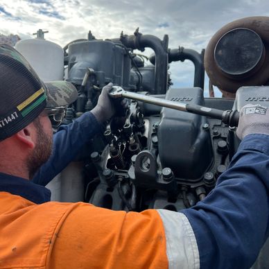 A mechanic in an orange and blue uniform repairs an engine outdoors.