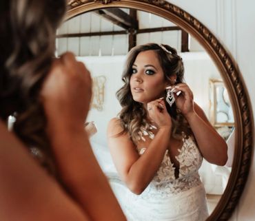 Bride adjusting earrings in front of a mirror, wearing a lace wedding dress.