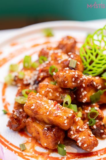 Spicy sesame-coated fried tofu garnished with spring onions and a decorative green leaf.