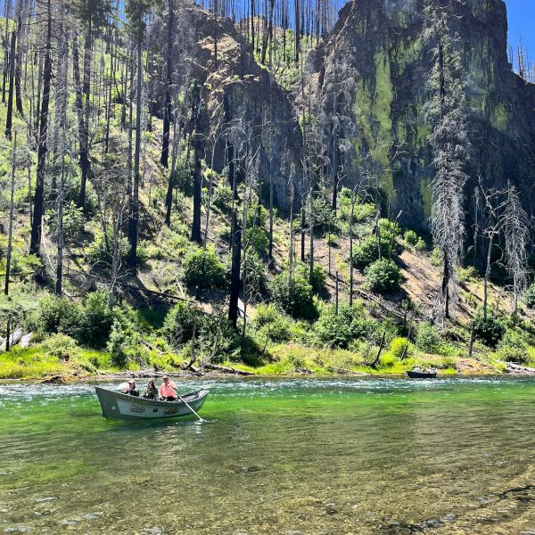 People fishing in boats on a clear river surrounded by forest and cliffs.