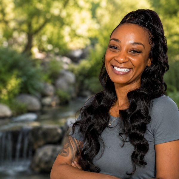 Smiling woman with long black hair standing outdoors near a stream.
