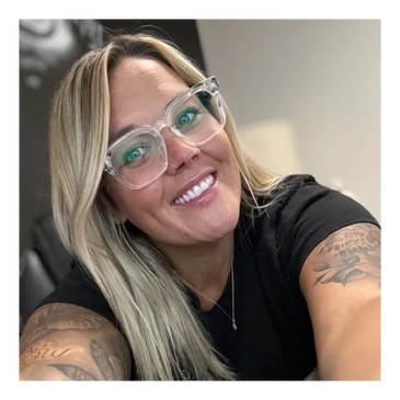 Smiling woman with blonde hair, clear glasses, and tattoos on both arms taking a selfie.