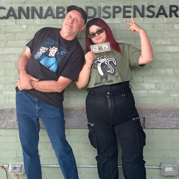 Two people posing happily in front of a cannabis dispensary sign.