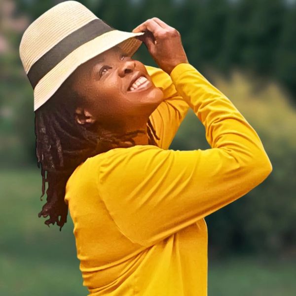 Smiling woman in yellow shirt adjusting her white hat outdoors.