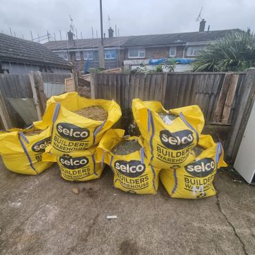 Yellow bags of builders' materials stacked outdoors near a wooden fence.