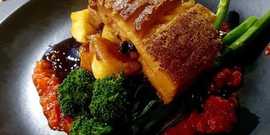 Crispy pork belly with vegetables and red sauce on a black plate.