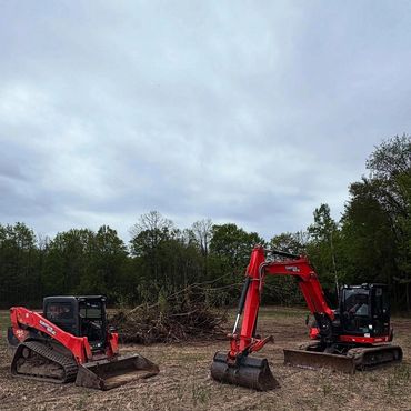 Two red excavators parked on a cleared field near a pile of branches.
