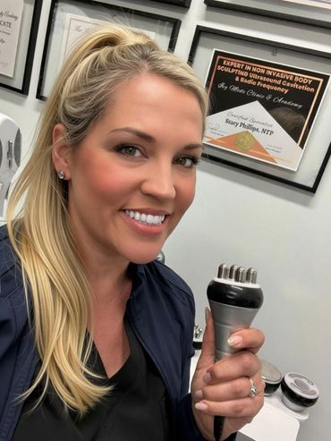 A smiling woman holding a body sculpting device in a clinic setting.