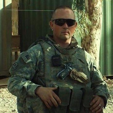 Soldier in full combat gear wearing sunglasses, standing outdoors.