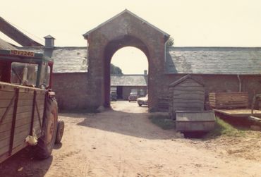 Farmyard entrance with tractor and rustic buildings under bright light.