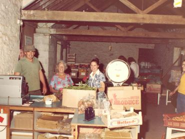People smiling at a rustic market stall with fresh produce and a vintage scale.