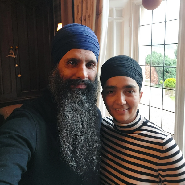 Two people wearing turbans pose indoors near a window with a garden view.
