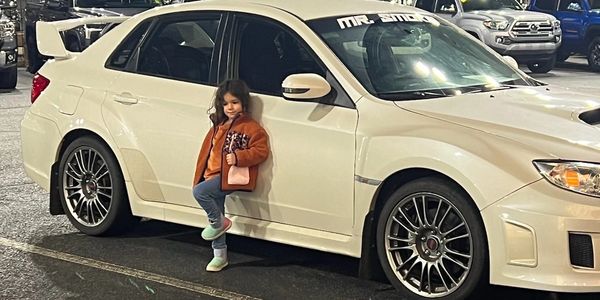A young girl leans against a white sports car in a parking lot at night.