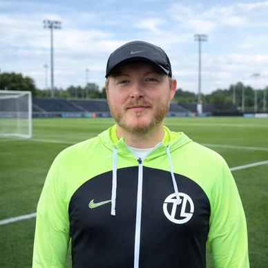 Man in a sports jacket and cap on a soccer field.