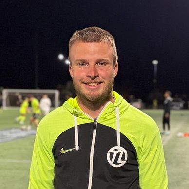 Young man in a neon green and black sports jacket smiling on a soccer field at night.