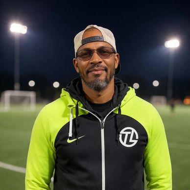 Man in a neon and black jacket with a cap stands on a lit soccer field at night.