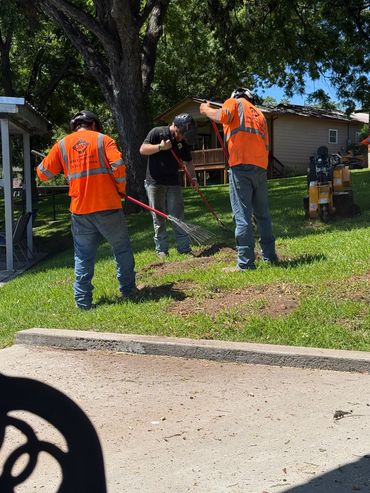 Three workers wearing safety gear raking grass near a tree on a sunny day.