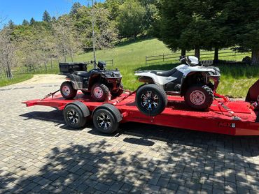 Two ATVs secured on a red trailer parked on a stone driveway.