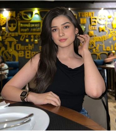 Young Karachi Escorts posing at a restaurant table with artistic yellow wall decor.