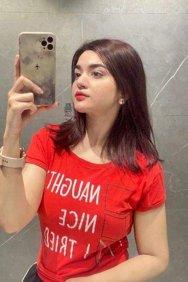Young escorts Karachi in red shirt taking a mirror selfie with her phone.