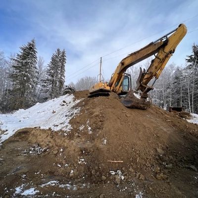 Excavator digging dirt on a snowy hill with a forest backdrop.